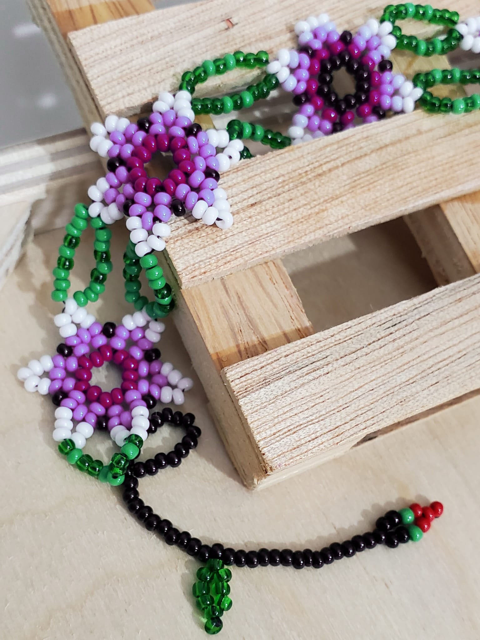 Zoom of Bracelet with five purple and white flowers connected with green petals and small cherry at the end. All made with glass beads.