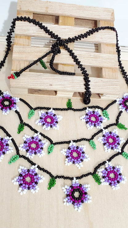 Glass beaded necklace with black zig zag beaded chain, that has 12 purple tone with white flowers in three chains on the front with green leaves in between the flowers. On the end of the chain there is a small beaded cherry. All made with glass beads.