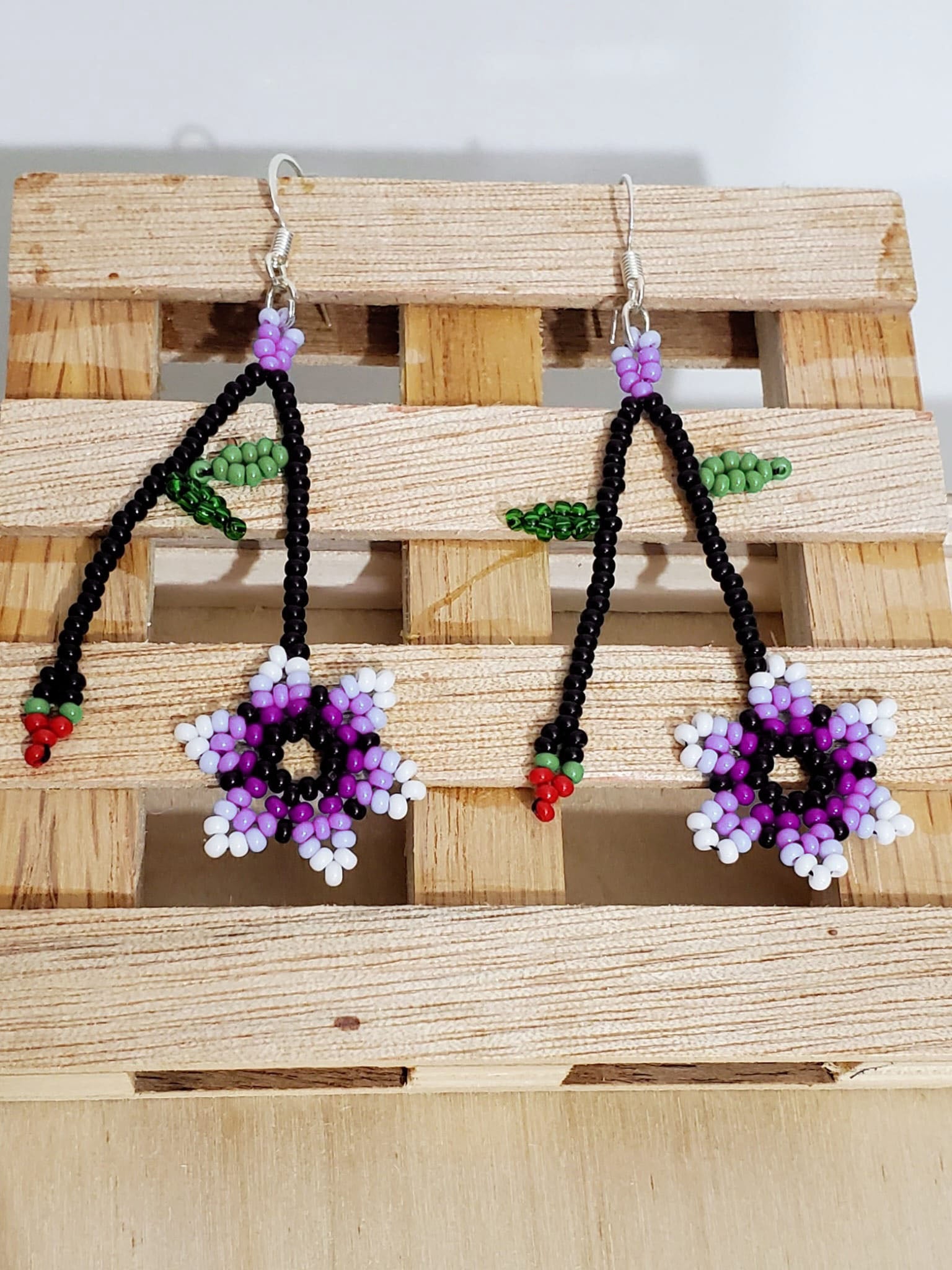 Pair of beaded earrings long with black beads, with two chains, purple and white flower at one end and small cherry with petals on the other. Earring has hook metal.